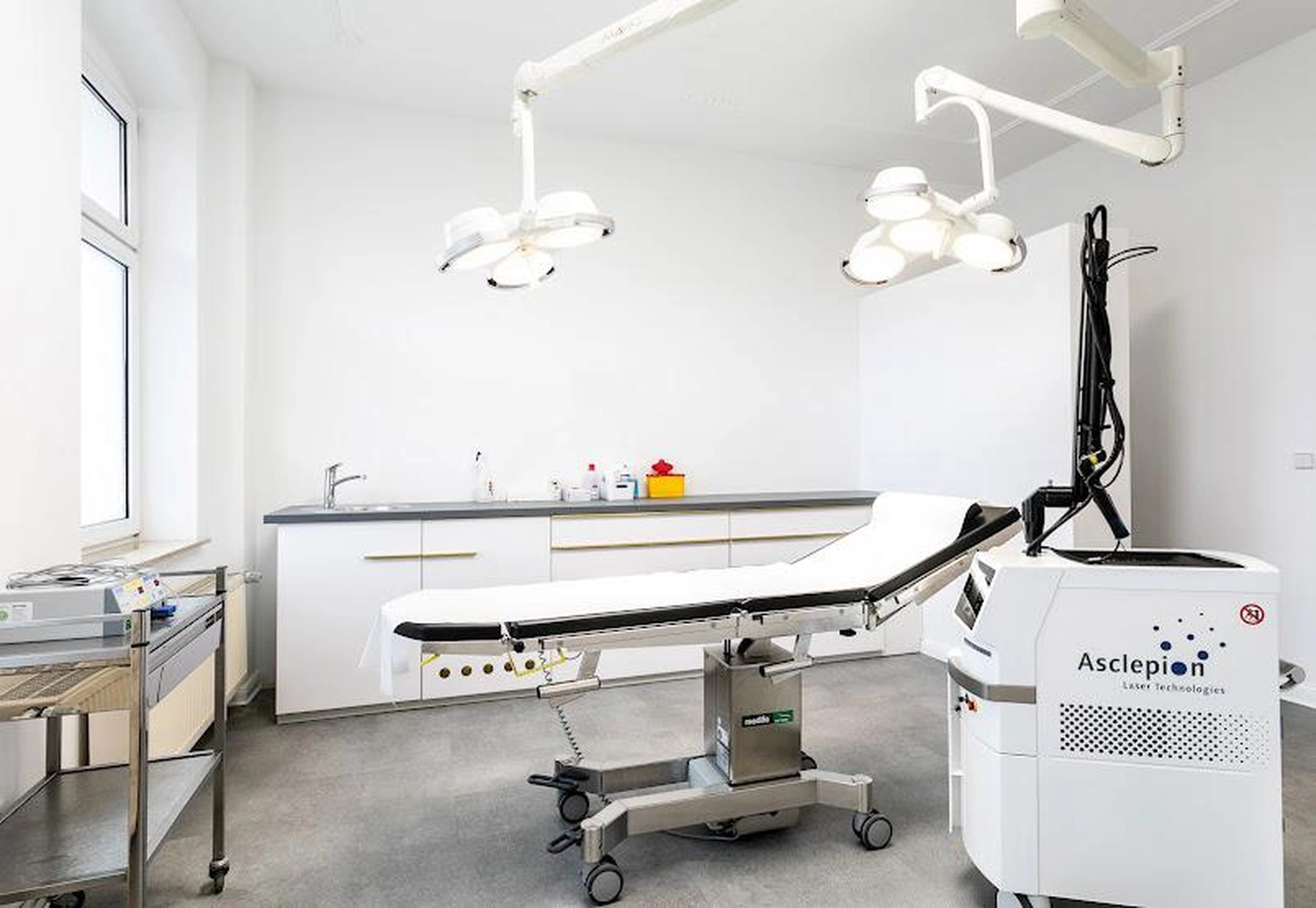 Flink Mediclean - Hygienische Reinigung für medizinische & therapeutische Einrichtungen - A bright, modern medical treatment room featuring a white adjustable examination table in the center under surgical lights. To the right stands an Asclepion laser machine, while a stainless steel cart and a long white counter with a sink line the walls.