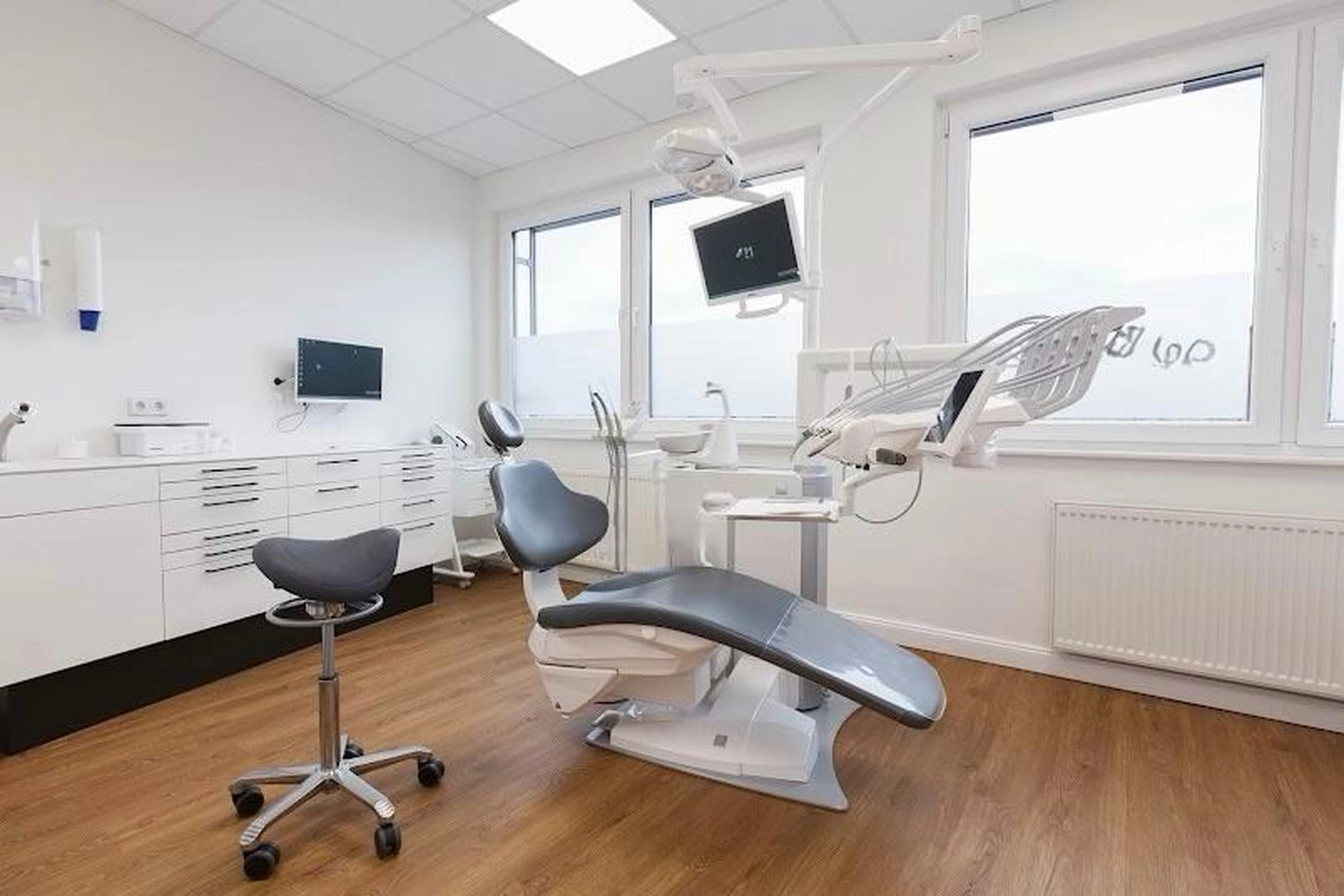 Flink Mediclean - Hygienische Reinigung für medizinische & therapeutische Einrichtungen - Ein sauberer, moderner Zahnarztbehandlungsraum mit einem zentralen grauen Liegestuhl, einem rollbaren Sattelhocker und medizinischen Geräten vor weißen Wänden und hellem Holzboden.