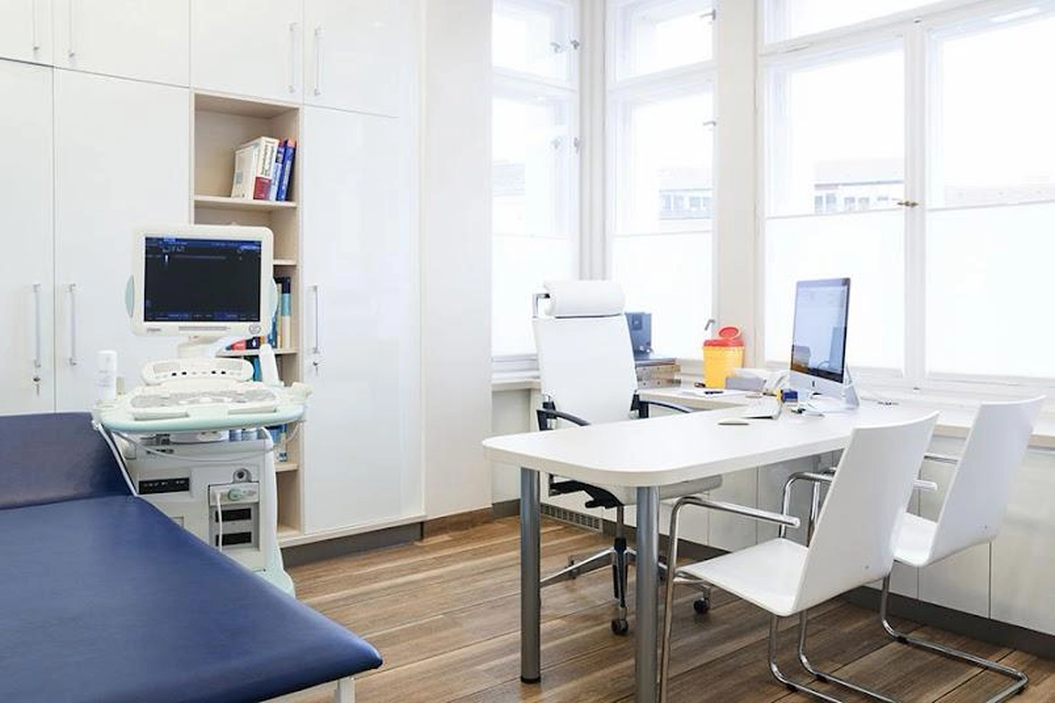 Flink Mediclean - Hygienische Reinigung für medizinische & therapeutische Einrichtungen - Ein organisierter, moderner Untersuchungsraum mit einer blauen Untersuchungsliege, einem Ultraschallgerät und einem weißen Schreibtisch mit mehreren Stühlen.