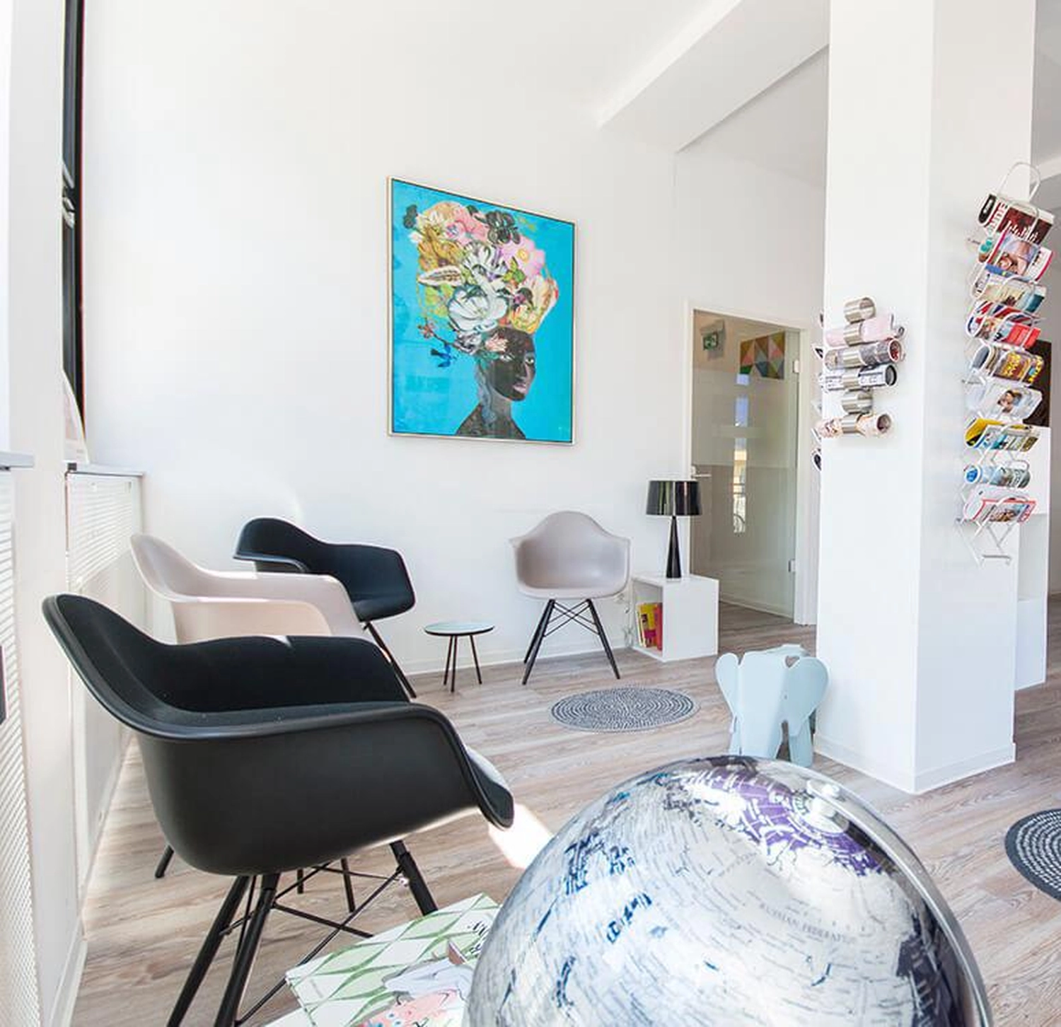 Flink Mediclean - Hygienische Reinigung für medizinische & therapeutische Einrichtungen - Ein helles, modernes Wartezimmer mit schwarz-weißen Sesseln im Eames-Stil, einem großen, farbenfrohen Porträt einer Frau mit floralem Kopfschmuck und einem dekorativen Globus im Vordergrund.