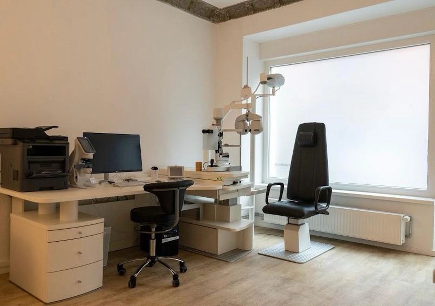 Flink Mediclean - Hygienische Reinigung für medizinische & therapeutische Einrichtungen - Ein Augenuntersuchungsraum mit einem Schreibtisch, auf dem ein Computer und medizinische Geräte stehen, einem verstellbaren Untersuchungsstuhl und einem Phoropter, der an einem großen Fenster positioniert ist.