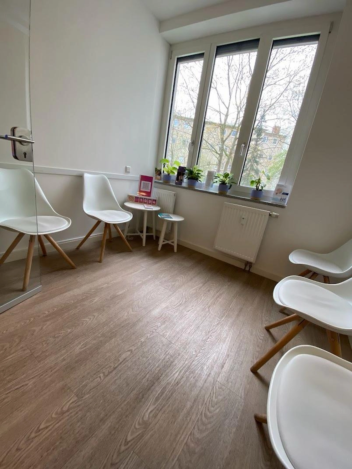 Flink Mediclean - Hygienische Reinigung für medizinische & therapeutische Einrichtungen - Ein helles Wartezimmer mit mehreren weißen, modernen Stühlen, einem hellen Holzboden und großen Fenstern mit Blick auf Bäume.