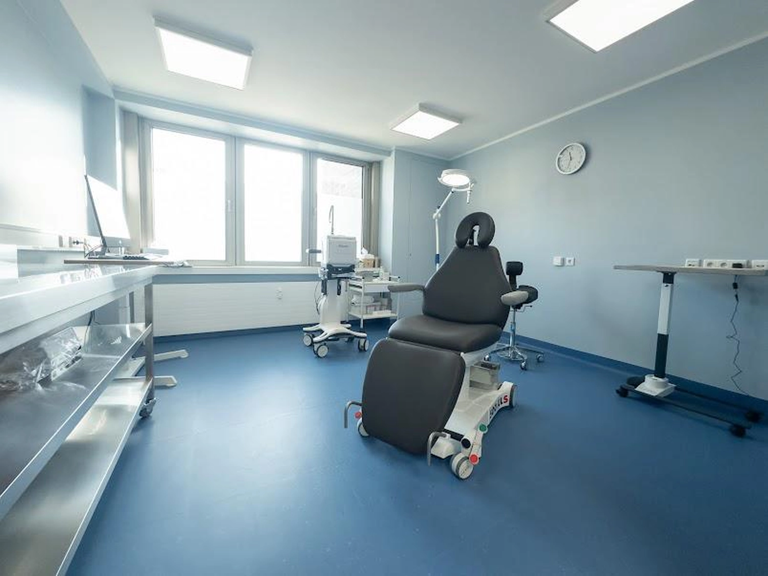 Flink Mediclean - Hygienische Reinigung für medizinische & therapeutische Einrichtungen - Das Bild zeigt einen modernen, sauberen Untersuchungs- oder Behandlungsraum mit blauem Boden und hellblauen Wänden. In der Mitte befindet sich ein spezieller, schwarzer, verstellbarer Behandlungsstuhl auf Rollen, umgeben von verschiedenen Geräten, darunter ein mobiler Arbeitsplatz, eine Wanduhr und eine helle Deckenbeleuchtung.