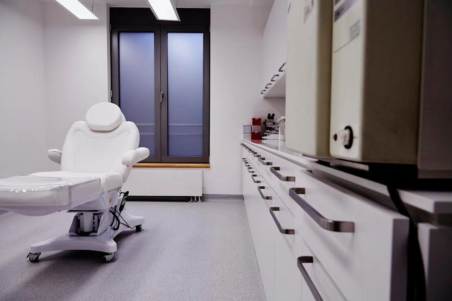 Flink Mediclean - Hygienische Reinigung für medizinische & therapeutische Einrichtungen - Ein makellos sauberer, moderner Behandlungsraum für medizinische oder ästhetische Behandlungen mit einem weißen hydraulischen Behandlungsstuhl, eleganten weißen Schränken mit langen Metallgriffen und großen Milchglasfenstern.
